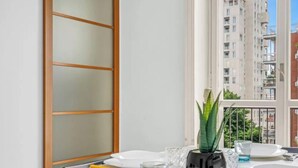 Una tavola imbandita con piatti, posate e bicchieri, una pianta in vaso e vista su edifici da una finestra.