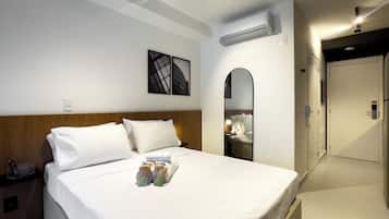 A hotel room with a bed, bedside table, nightstand, wall-mounted air conditioner, and a door leading to another room.