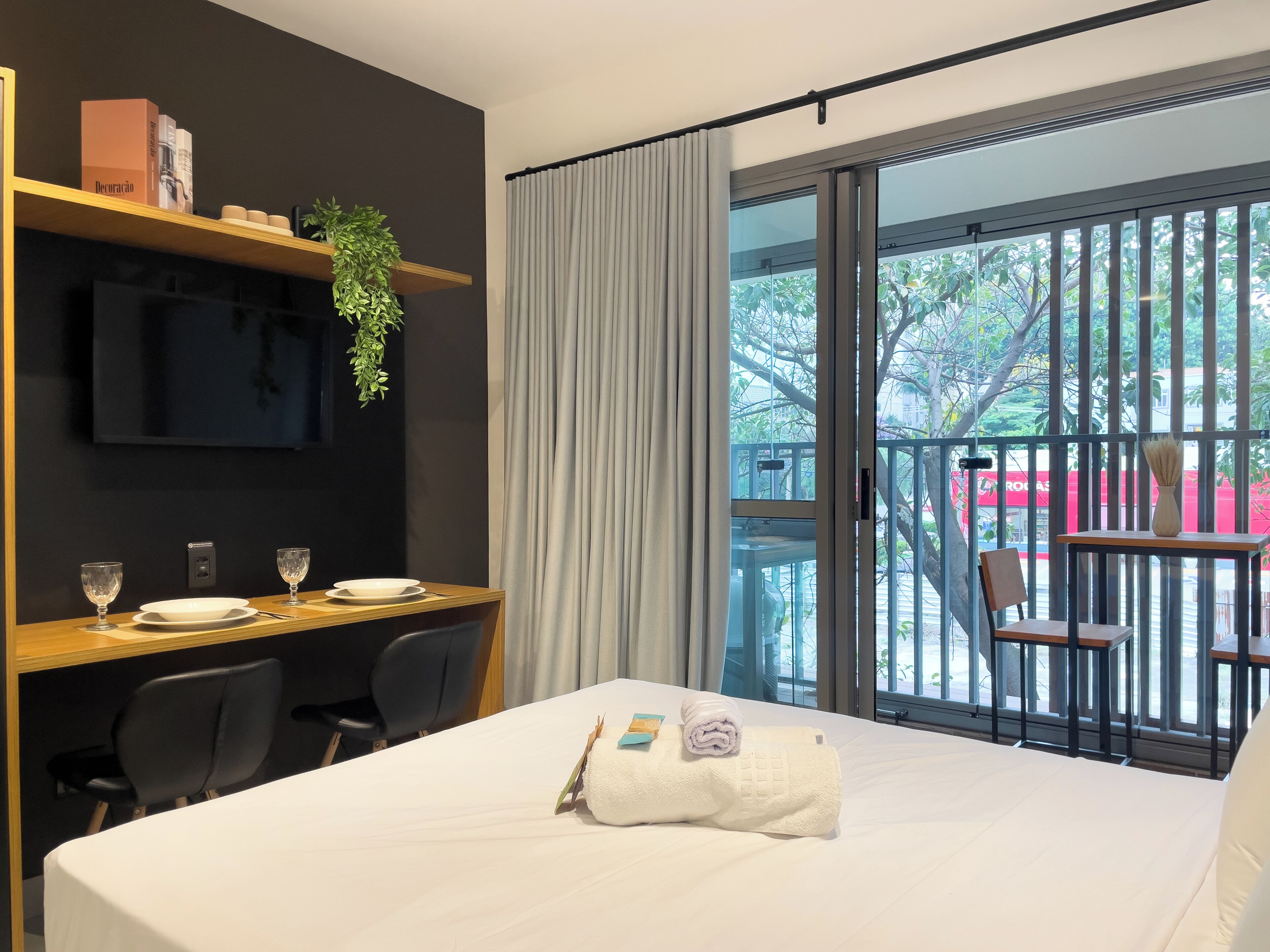 A hotel room with a bed, a desk with two chairs, a TV mounted on the wall, and a balcony with a chair and railing.