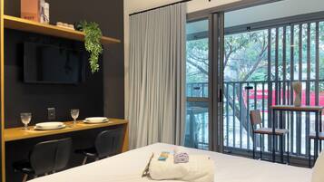A hotel room with a bed, a desk with two chairs, a TV mounted on the wall, and a balcony with a chair and railing.