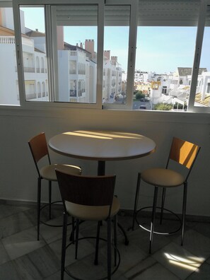 Ein Zimmer mit rundem Tisch und vier Stühlen, großen Fenstern mit Blick auf Gebäude und einem Fliesenboden.