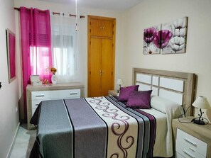 Ein Schlafzimmer mit einem Bett, einem Nachttisch, einem Holzschrank, einem Fenster mit rosa Vorhängen und einem floralen Bild an der Wand.