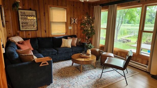 A cozy living room with a dark blue sectional sofa, a wooden coffee table, and a large window offering a view of a garden. - Lakefront Cabin - Paddleboat, Paddleboards & Fishing Dock Included. Pet friendly (Lindstrom)