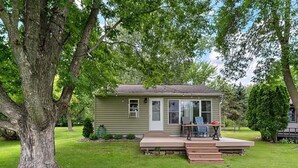 A small single-story house with a front porch, a lawn, and a large tree in the foreground. - Lakefront Cabin - Paddleboat, Paddleboards & Fishing Dock Included. Pet friendly (Lindstrom)