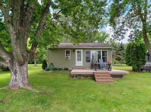 A small single-story house with a front porch, a lawn, and a large tree in the foreground. - Lakefront Cabin - Paddleboat, Paddleboards & Fishing Dock Included. Pet friendly (Lindstrom)