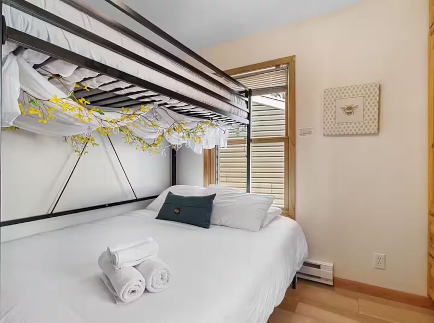 A bedroom with a bunk bed, a single bed, a window with blinds, a wall-mounted heater, and a framed picture.