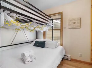 A bedroom with a bunk bed, a single bed, a window with blinds, a wall-mounted heater, and a framed picture.