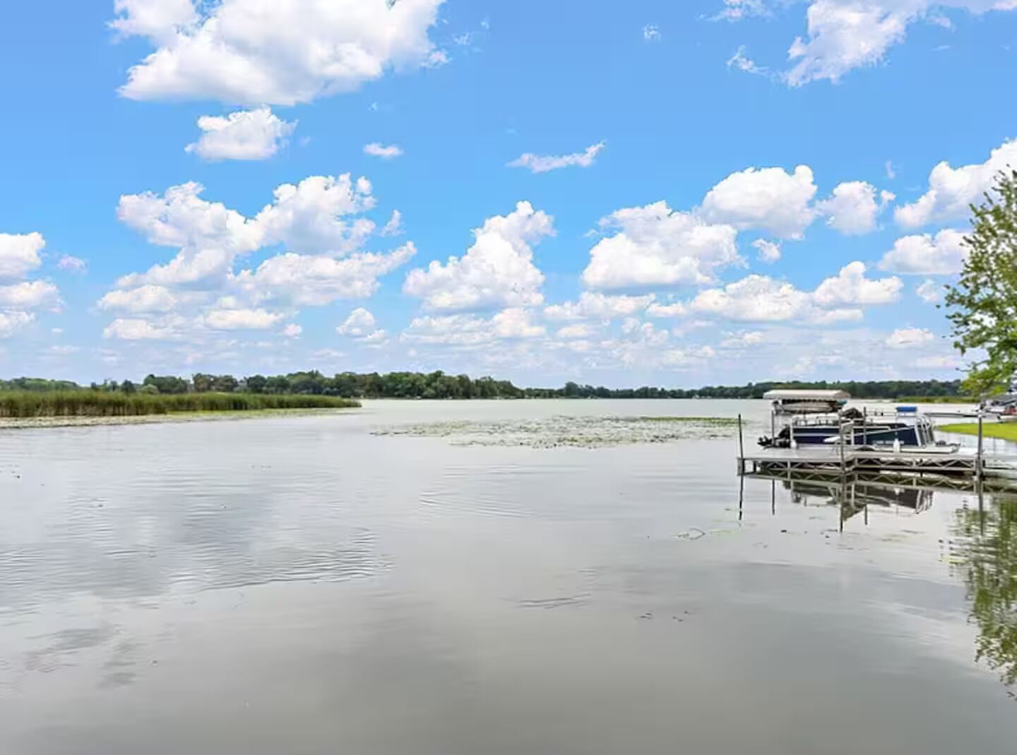 A calm lake with a dock and a boat, surrounded by greenery and a clear sky.
