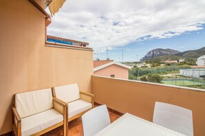 Un balcon avec deux chaises blanches dotées de coussins et une table, offrant une vue sur un paysage montagneux et un terrain de sport.