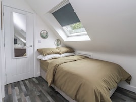 Un dormitorio con techo inclinado, tragaluz, cama con ropa de cama color beige, puerta blanca y un espejo.