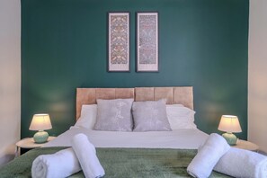 Un lit bien fait, avec des oreillers, une couverture verte et des serviettes enroulées posées sur la tête de lit. Deux cadres avec des photos sont accrochés au mur au-dessus du lit.