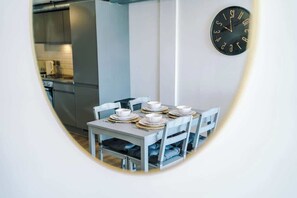 A dining area with a table set for four, a clock on the wall, and a kitchen in the background.