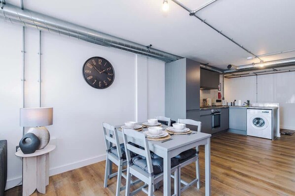 A modern kitchen with a built-in washing machine, a dining area with a table set for four, and a wall-mounted clock.