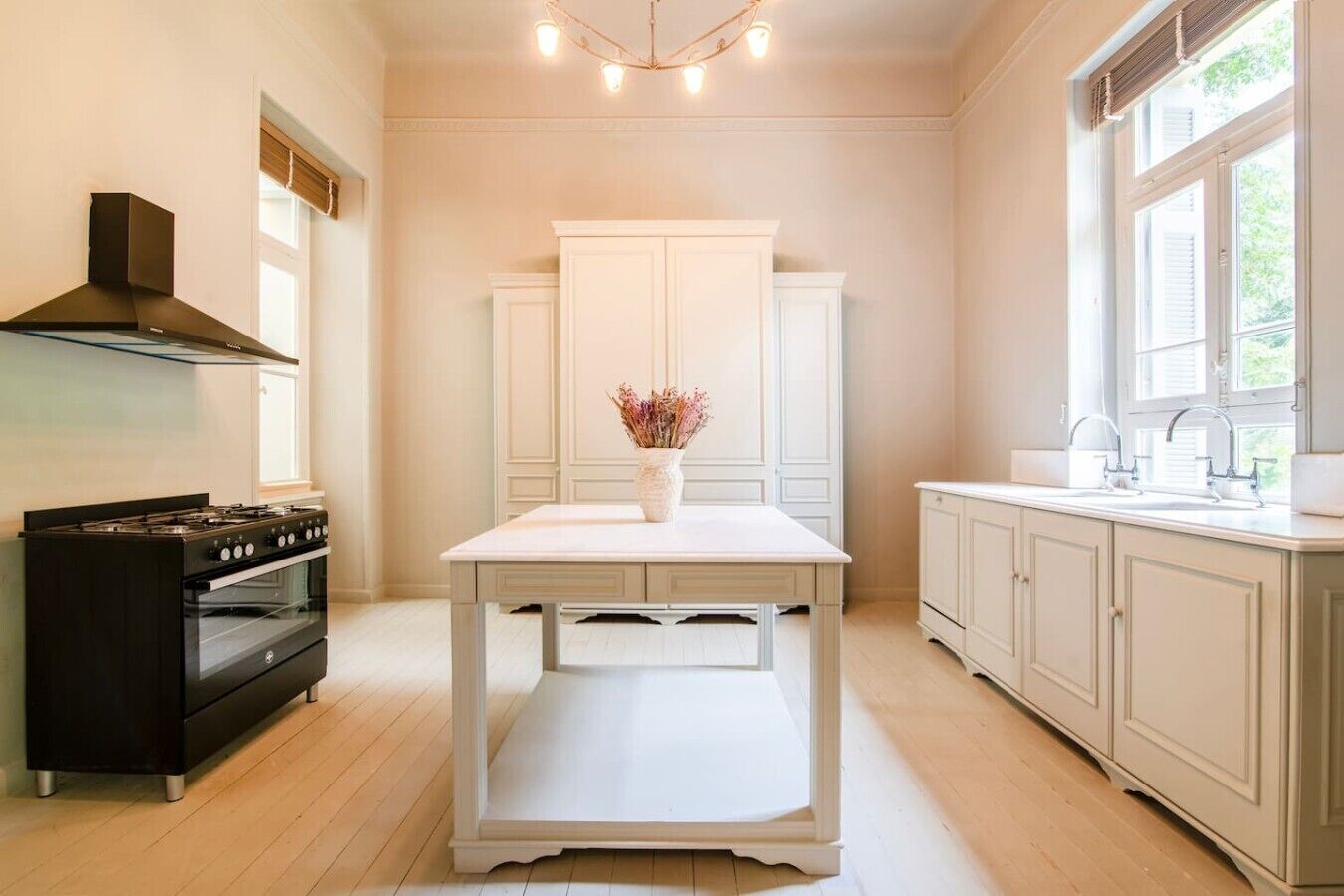 A kitchen with a stove, oven, white cabinets, a table with a vase, and a window.