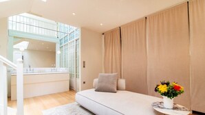 Une chambre moderne avec un canapé-lit, une petite table ornée d’un vase de fleurs, une salle de bain avec une paroi en verre et un escalier.