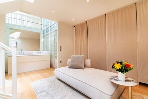 Une chambre moderne avec un canapé-lit, une petite table ornée d’un vase de fleurs, une salle de bain avec une paroi en verre et un escalier.