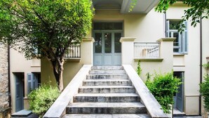 A set of stone steps leading to a door with a metal gate, surrounded by greenery and a building with a balcony.