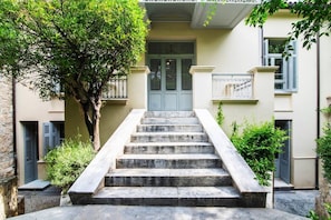 A set of stone steps leading to a door with a metal gate, surrounded by greenery and a building with a balcony.