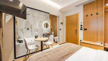 Une chambre d’hôtel moderne équipée d’un lit, d’une salle de bain avec douche, d’un meuble-lavabo et d’un coin salon avec des fauteuils.