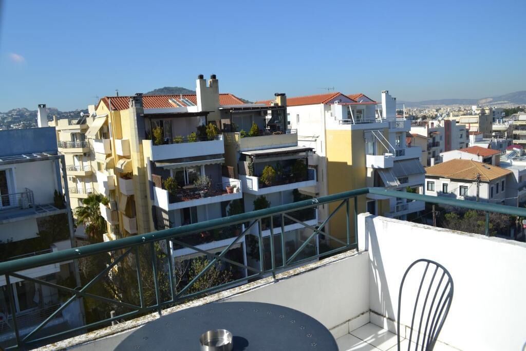 Un balcone con tavolo e sedie, vista su una zona residenziale con edifici e cielo sereno.