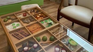 Hotellihuone, jossa on sänky, puinen yöpöytä lampun kanssa, tuoli ja tarjoilupöytä, jossa on erilaisia esineitä.