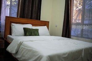 A neatly made bed with white bedding and a single green pillow, a wooden headboard, and a window with curtains.