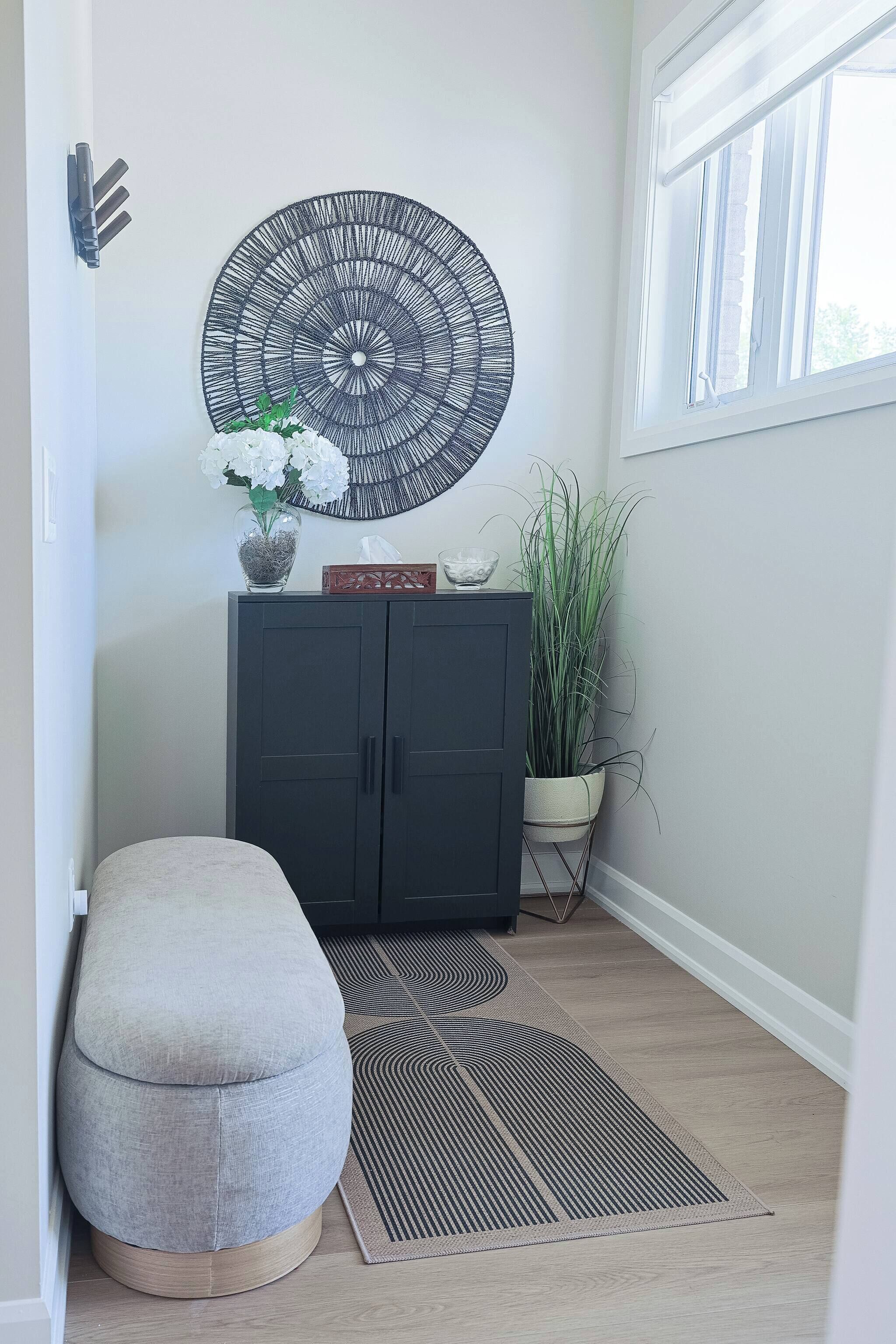A hallway with a grey ottoman, a black cabinet with decorative items, a large circular wall art, and a potted plant.