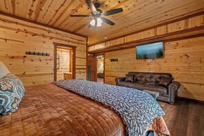 A log cabin bedroom with a bed, a brown leather couch, a ceiling fan, and a window with a view of trees. - 8-Bedroom Smoky Mountain Cabin with Indoor Pool, Hot Tub & Game Room (Sevierville)
