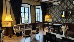 Uma área de refeições com mesa de madeira, cadeiras e um banco. Há duas janelas com cortinas, uma parede com papel de parede estampado e um lustre.