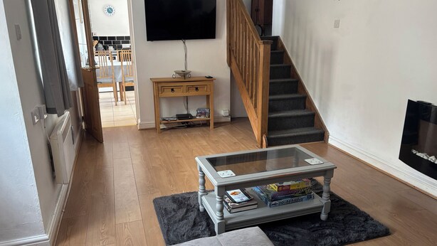 A living room with a sofa, a coffee table with books, a TV, a staircase, and a dining area in the background. - Tor Cefn (Kidwelly)
