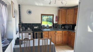 A kitchen with wooden cabinets, a dining table with chairs, a microwave, and a window with a view of greenery. - Tor Cefn (Kidwelly)