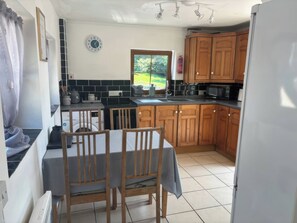 A kitchen with wooden cabinets, a dining table with chairs, a microwave, and a window with a view of greenery. - Tor Cefn (Kidwelly)
