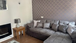 A living room with a grey sofa, a small wooden side table, a fireplace, and a patterned wallpaper. - Tor Cefn (Kidwelly)