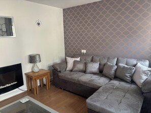 A living room with a grey sofa, a small wooden side table, a fireplace, and a patterned wallpaper. - Tor Cefn (Kidwelly)