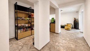 A modern interior with a kitchen area featuring a wooden pantry shelf stocked with food items, a seating area with a yellow chair, and a reception desk with a computer monitor. - BASE Achensee Balance N°01 – Garden (Achenkirch)