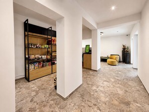 Ein modernes Interieur mit Küchenzeile, an der sich ein Vorratsschrank aus Holz mit Lebensmitteln, eine Sitzecke mit einem gelben Sessel und ein Empfangsschalter mit Computerbildschirm befinden.