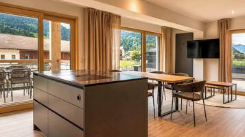 A modern kitchen with a central island, dining area with chairs, large windows with curtains, and a flat-screen TV mounted on the wall.