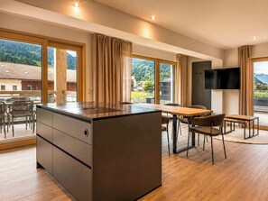A modern kitchen with a central island, dining area with chairs, large windows with curtains, and a flat-screen TV mounted on the wall. - BASE Achensee Freedom N°11 – Terrace (Achenkirch)