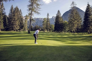 Eine Person spielt Golf auf einer gepflegten Golfanlage mit hohen Bäumen und Bergen im Hintergrund.