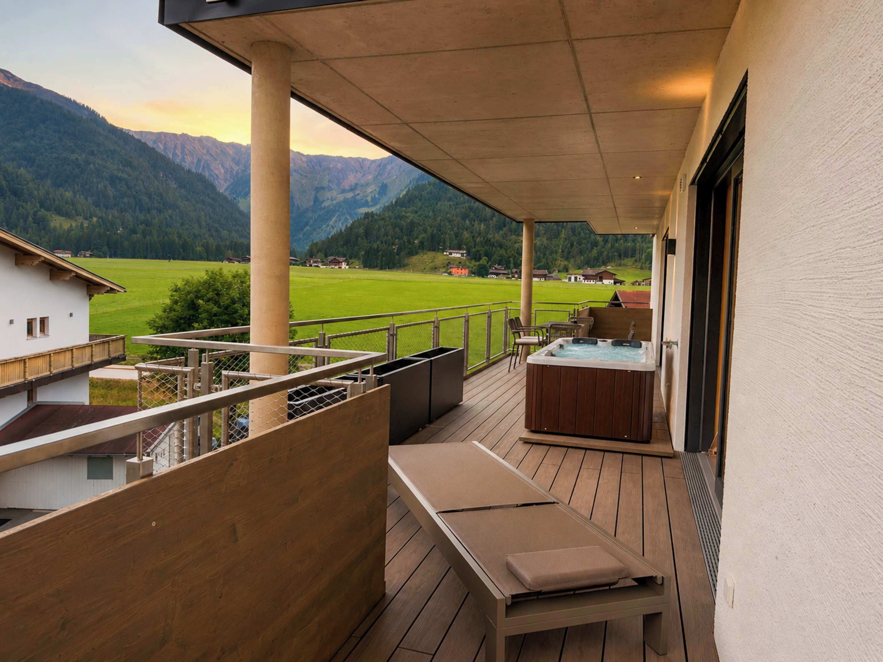 Eine Holzterrasse mit Whirlpool und Sitzgelegenheit mit Blick auf ein grünes Feld und Berge.