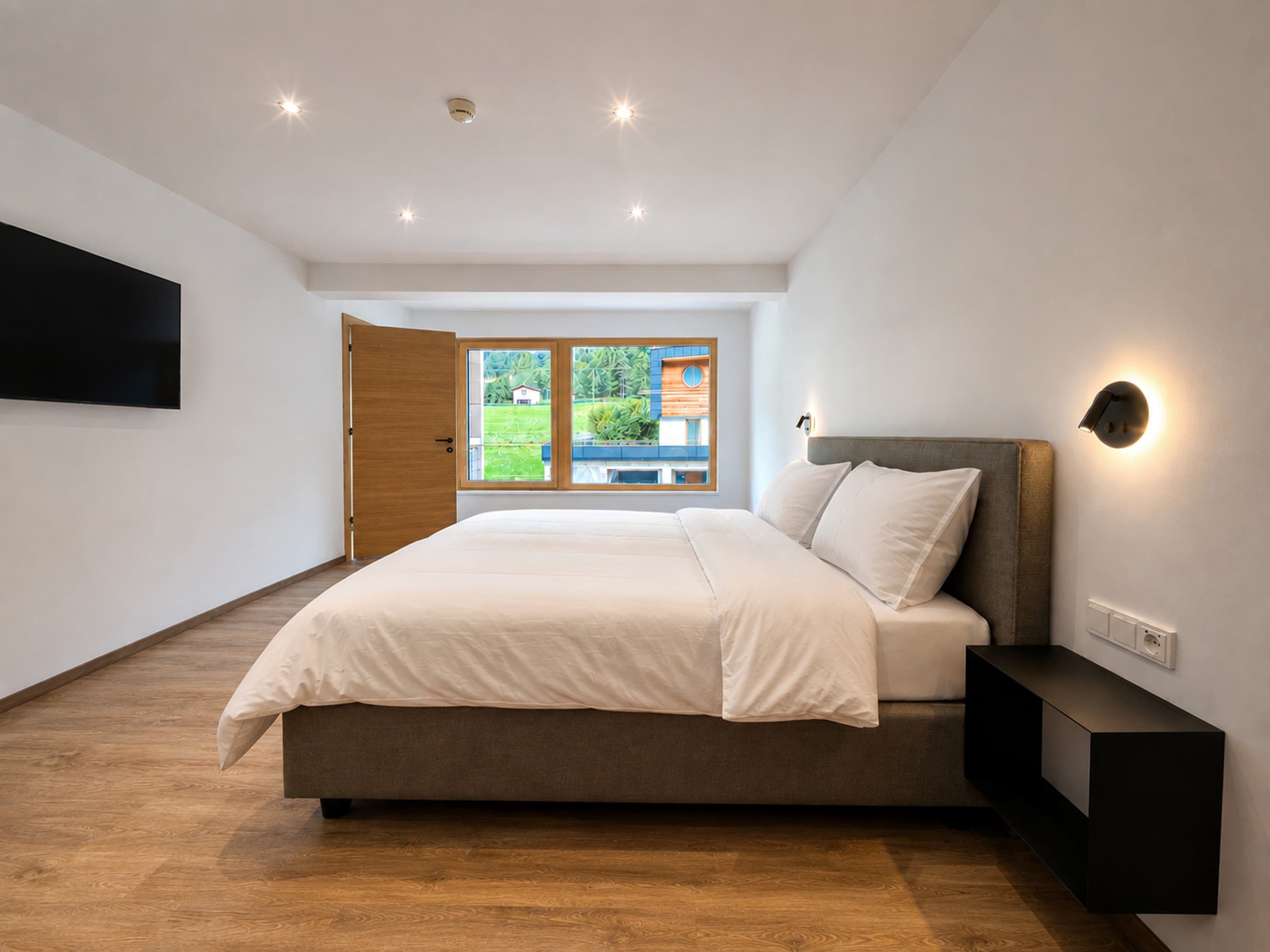 Une chambre moderne avec un grand lit, une table de chevet, un téléviseur fixé au mur et une fenêtre donnant sur un paysage verdoyant et des bâtiments.