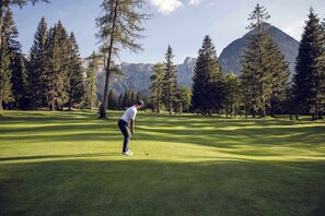 Una persona gioca a golf su un percorso ben curato, con alberi alti e montagne sullo sfondo.