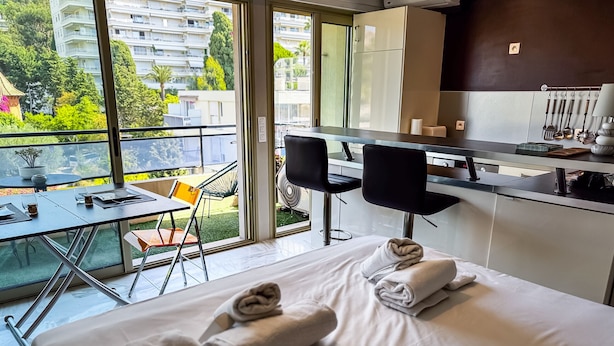A hotel room with a bed, a dining table, chairs, a kitchen area, and a balcony with a view of buildings and greenery. - Studio Terrasse Pointe Croisette A0b214 (Cannes)
