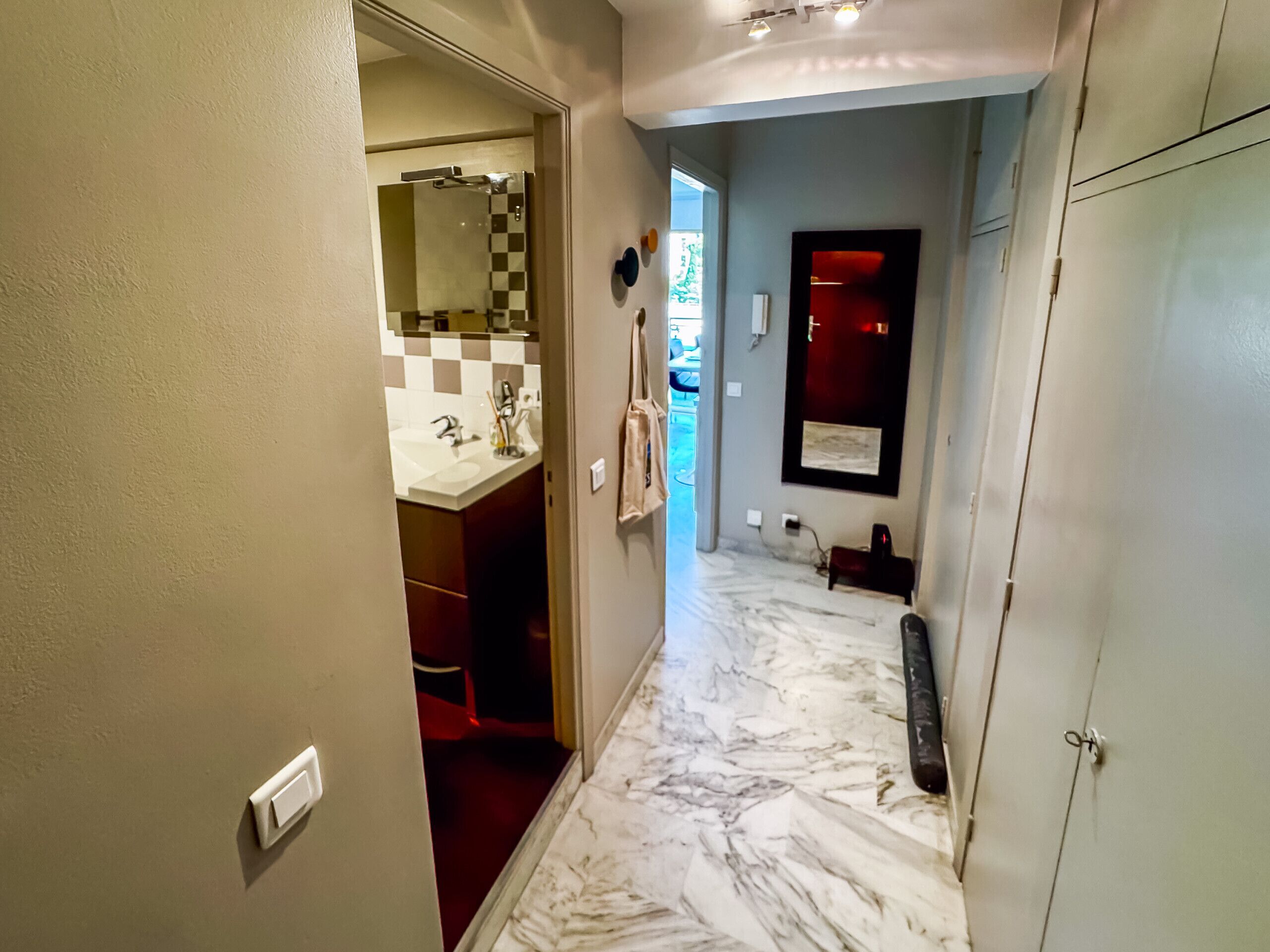 Un couloir doté d’un sol en marbre, d’un miroir, d’un mur rouge et d’une salle de bain visible à travers une porte ouverte.