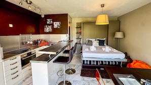 A modern hotel room with a kitchen area, a bed with a striped headboard, and a dining table set for two. - Studio Terrasse Pointe Croisette A0b214 (Cannes)