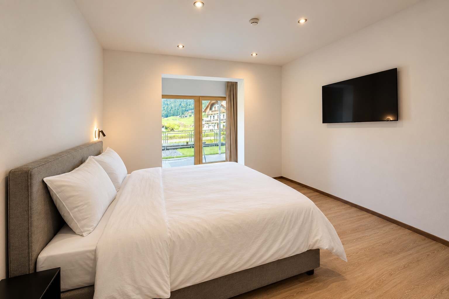 A modern bedroom with a large bed, a flat-screen TV mounted on the wall, and a view of a building through a glass door.