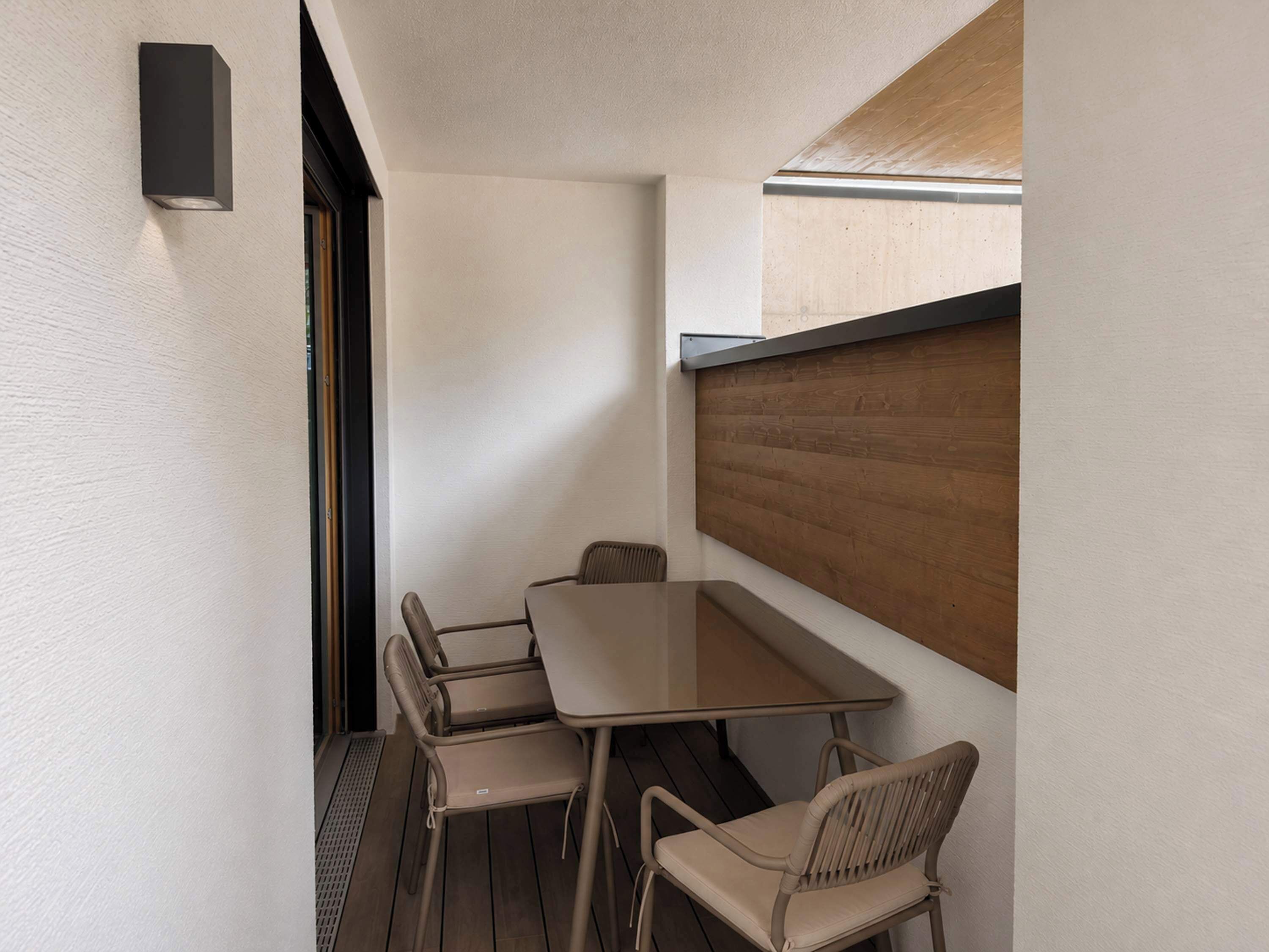 Un pequeño espacio al aire libre con comedor, una mesa y cuatro sillas, un muro de listones de madera y una lámpara en la pared.