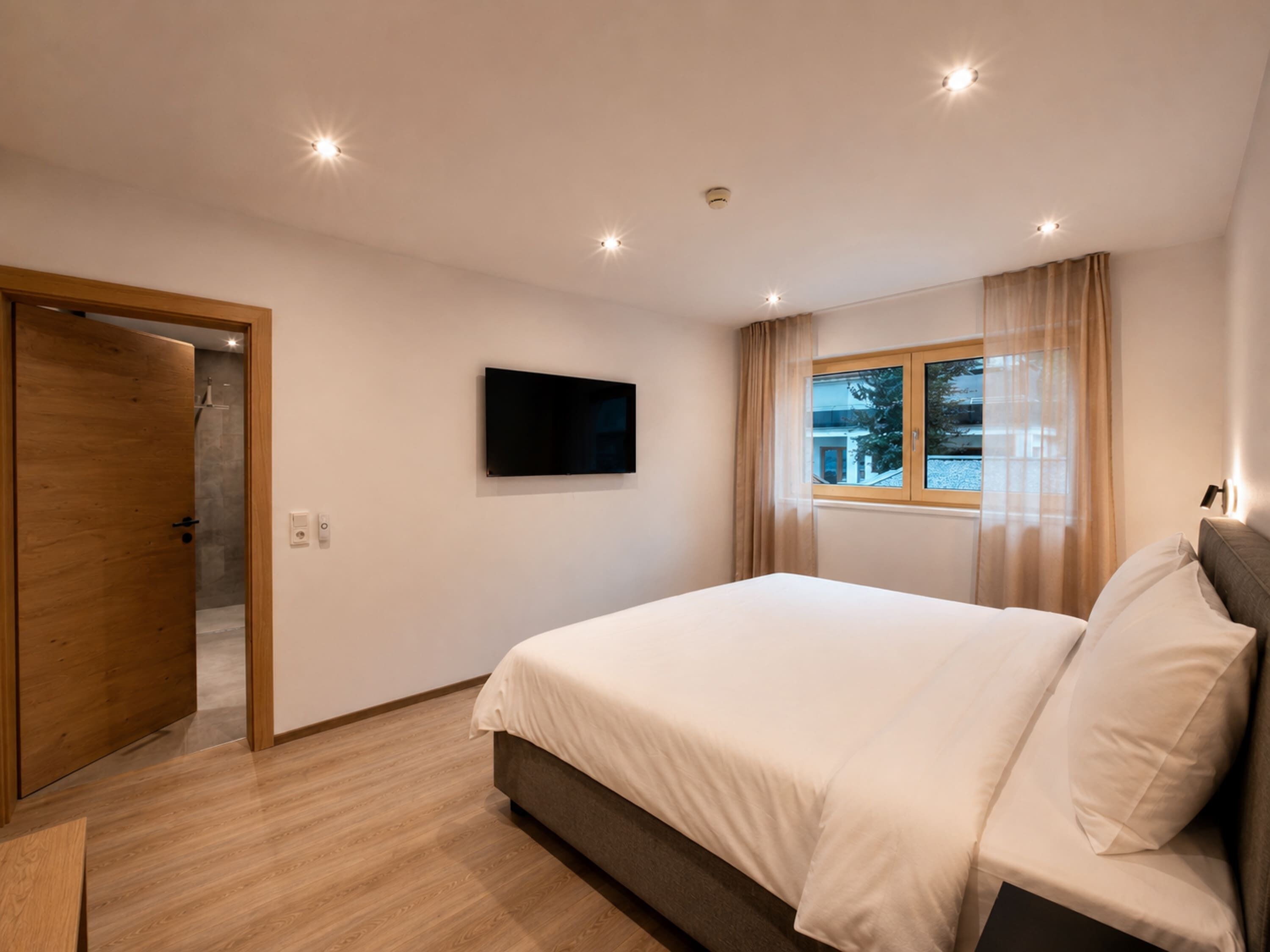 Een slaapkamer met een bed, een aan de muur bevestigde televisie, een raam met gordijnen en een deur naar een douchecel.
