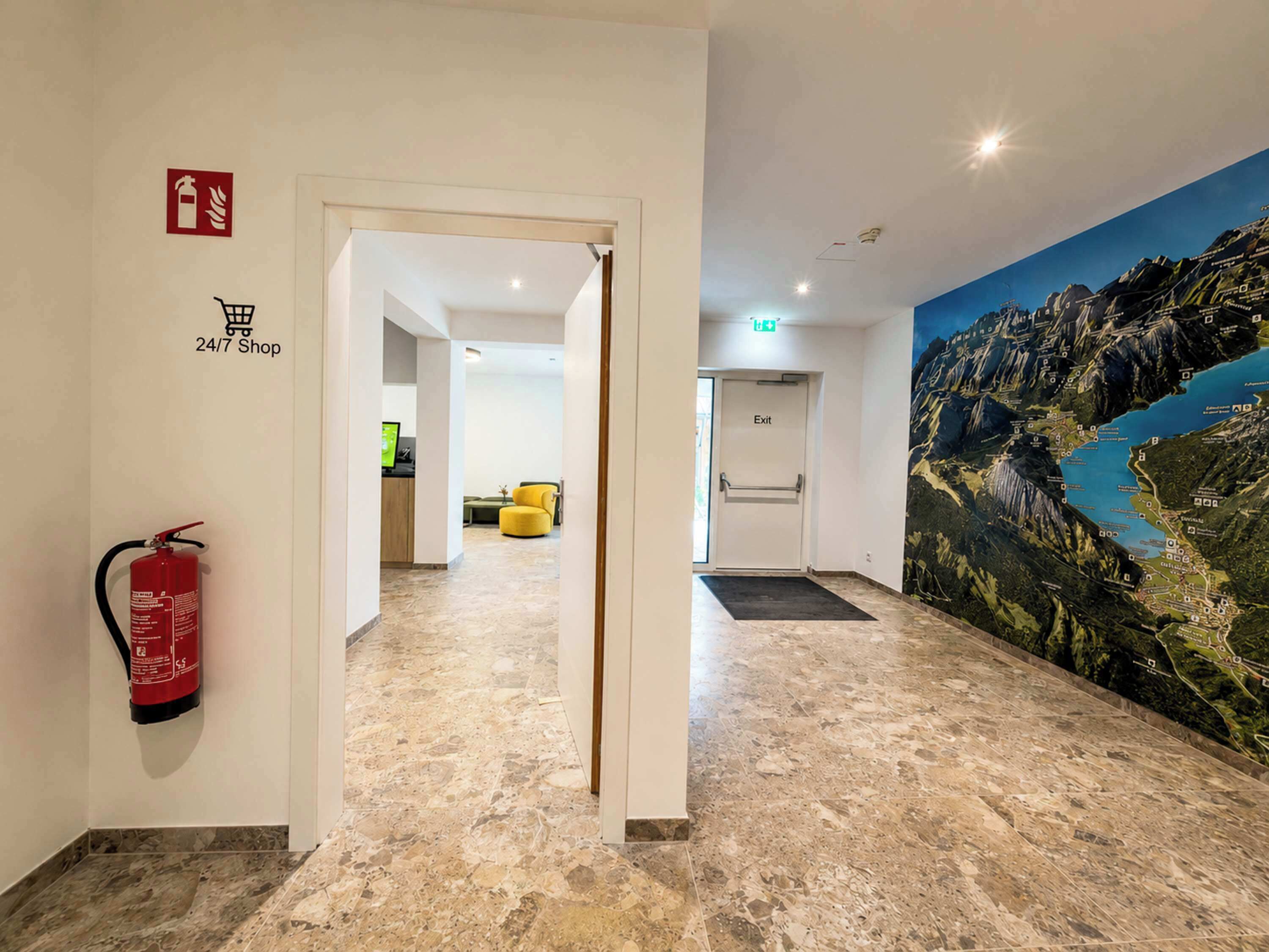 Un vestíbulo de hotel con un extintor, un letrero de tienda abierta las 24 horas y un mural de un paisaje montañoso.