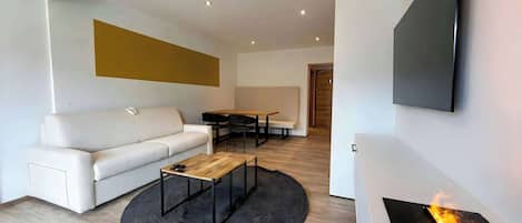 A modern living room with a white sofa, a coffee table, a dining area with a table and chairs, and a fireplace with a visible fire.